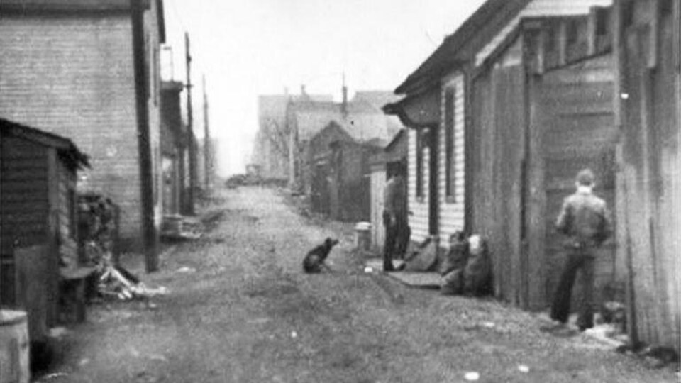 De petites maisons de bois le long d'une ruelle de terre où est assis un chien. Un homme rentre chez lui alors qu'un autre marche près des maisons du côté droit de la rue.