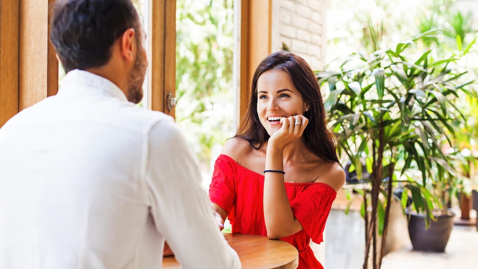 Une femme avec une robe rouge regarde un homme en souriant, tous les deux sont assis à une table.