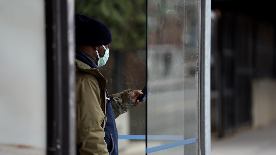 Un homme portant un masque regarde son cellulaire en attendant l'autobus. 