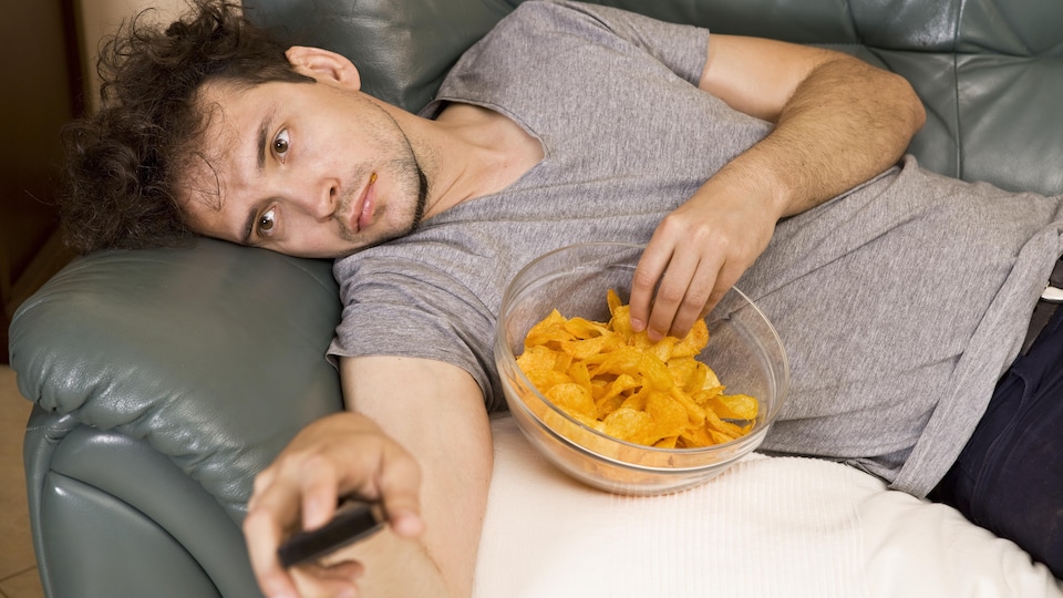 Un homme allongé sur un sofa mange des croustilles en regardant la télévision.