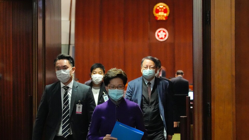 Carrie Lam sort d'une salle, suivi de quelques hommes. Les emblèmes de la Chine et de Hong Kong sont visibles en arrière-plan. 