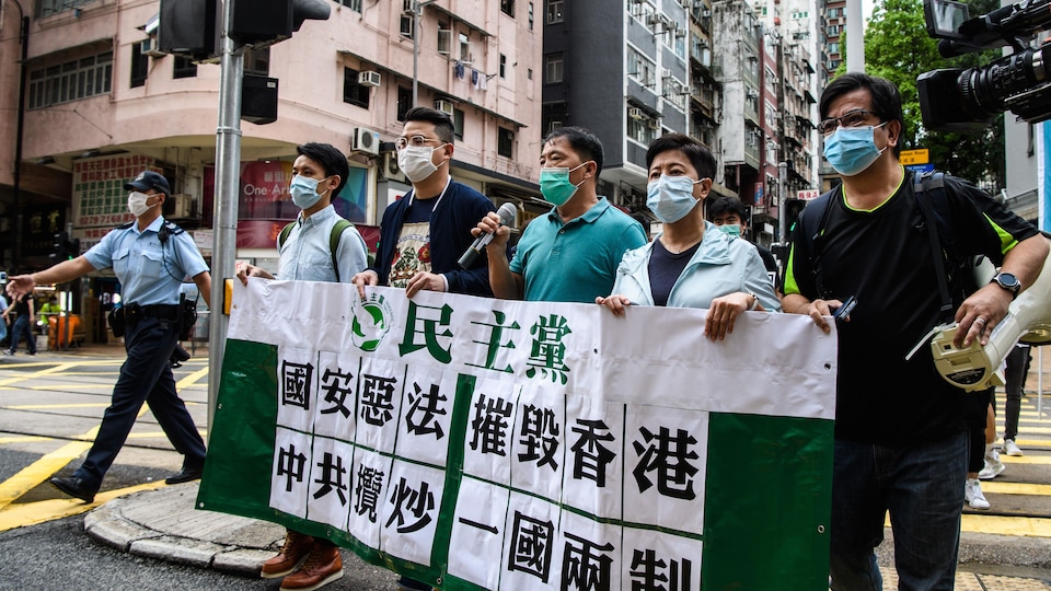 Cinq manifestants masqués marchent dans une rue en tenant une pancarte. L'un tient un micro, un autre, un mégaphone.