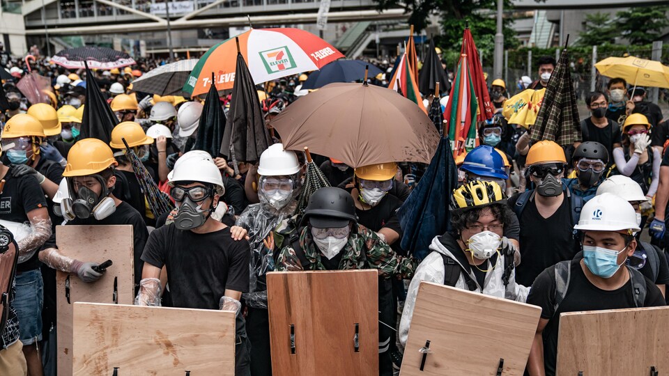 Une foule de manifestants masqués. Certains tiennent des boucliers faits de planches de bois; d'autres se couvrent d'un parapluie. La plupart d'entre eux portent un casque. 