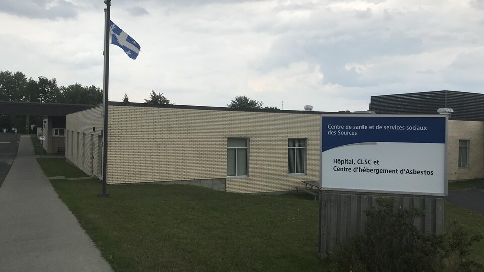 Un bâtiment, un drapeau du Québec et une enseigne en premier plan où il est indiqué, « Hôpital, CLSC et Centre d'hébergement d'Asbestos ». 