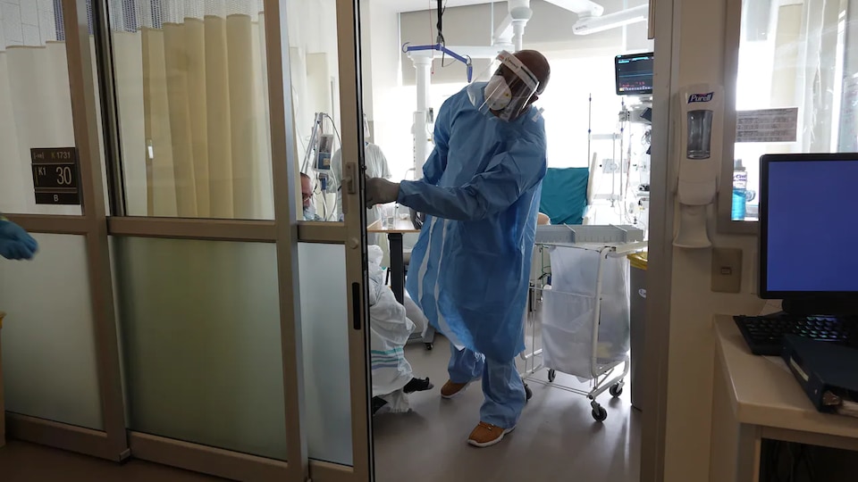 Un médecin en tenue de protection avec une visière et un masque se tient à l'entrée d'une chambre de soins intensifs d'un hôpital.