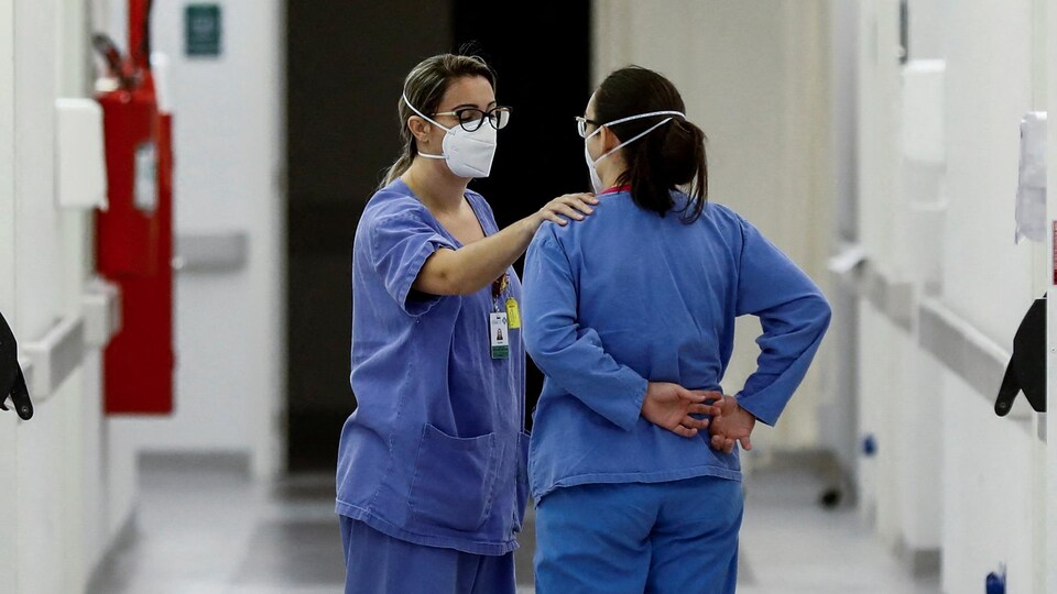Deux infirmières dans un couloir d'hôpital. Une met sa main sur l'épaule de l'autre.