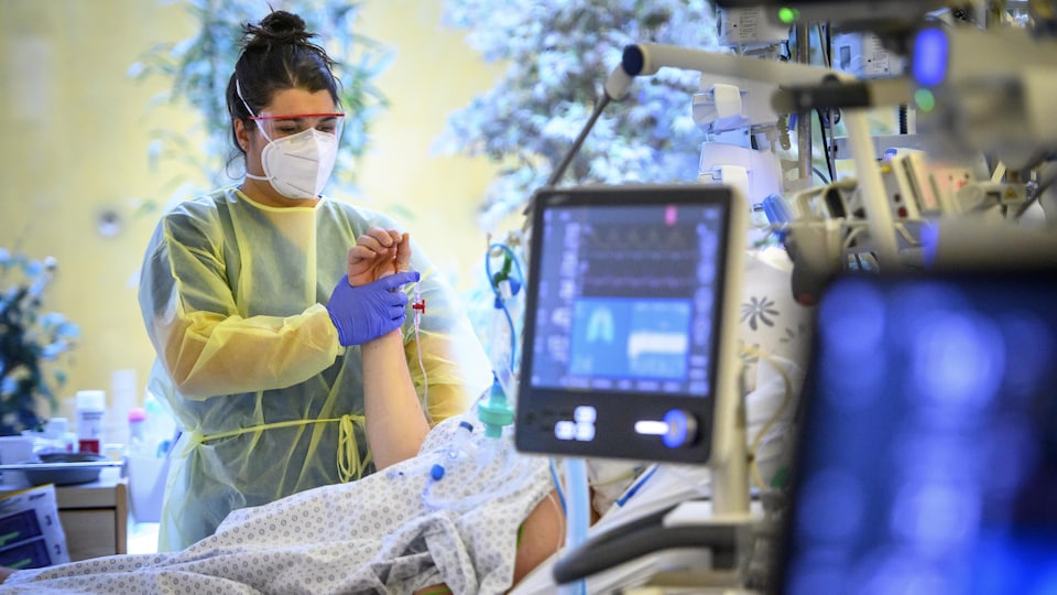 Une infirmière tient le bras d'une patiente couchée dans un lit d'hôpital aux soins intensifs. Le visage de la patiente est caché par un écran d'ordinateur.