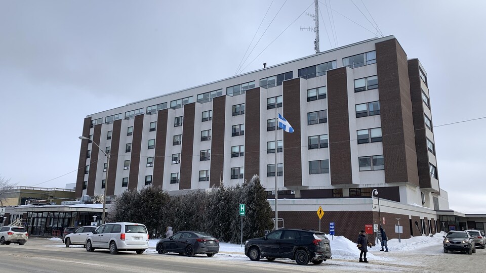 Des personnes marchent sur le trottoir devant le centre hospitalier. Des voitures sont aussi stationnées dans la rue.