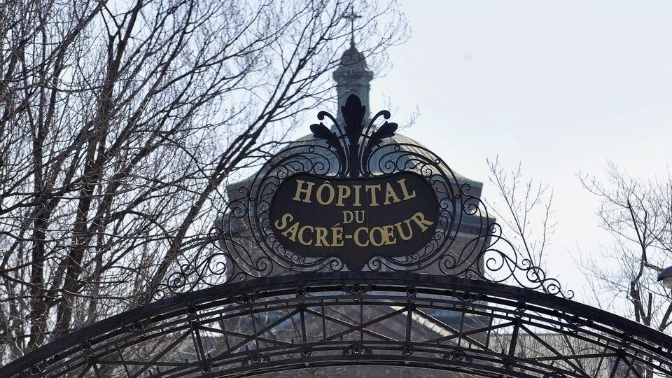 Un bâtiment, sur lequel se trouve une petite croix, est vu derrière une enseigne sur laquelle on peut lire « Hôpital du Sacré-Coeur ». 