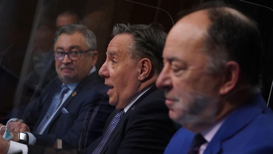 Horacio Arruda, François Legault et Christian Dubé sont assis côte à côte. Le premier ministre parle tandis que le Dr Arruda le regarde.