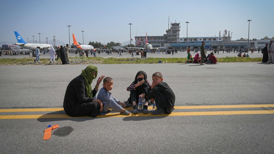 Des enfants et un adulte assis au milieu d'une route avec derrière des avions sur le tarmac d'un aéroport.