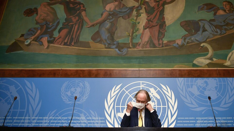 Un homme enlève son masque sur une tribune qui arbore le logo des nations unies et une énorme fresque.