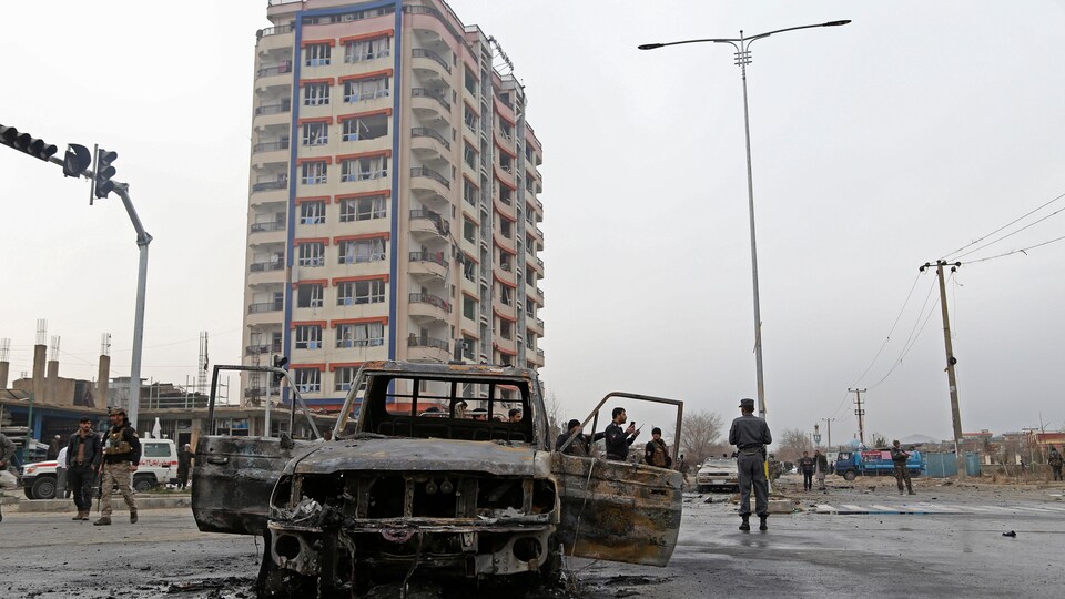 Des membres des forces de sécurité afghanes montent la garde sur le site de l'attentat, où l'on aperçoit une voiture entièrement calcinée. 