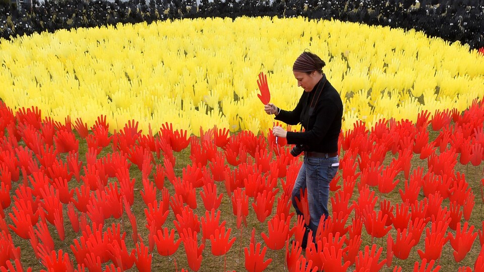 Une jeune femme est en train de planter dans le sol une main rouge. Autour d'elle, des dizaines de mains similaires sont déjà plantées. Elles sont de couleurs jaune (le soleil), rouge (la terre du désert) et noire (la couleur de leur peau).