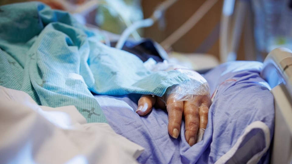 Un patient est dans un lit d'hôpital. On voit sa main en gros plan.