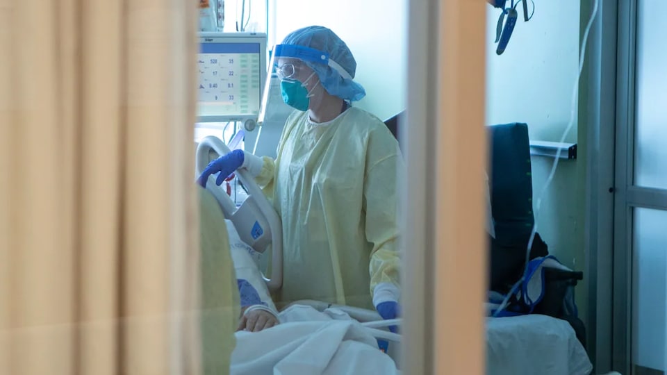 Une infirmière qui porte une jaquette jaune, des gants bleus, un bonnet bleu, un masque et une visière est au chevet d'un lit d'hôpital.