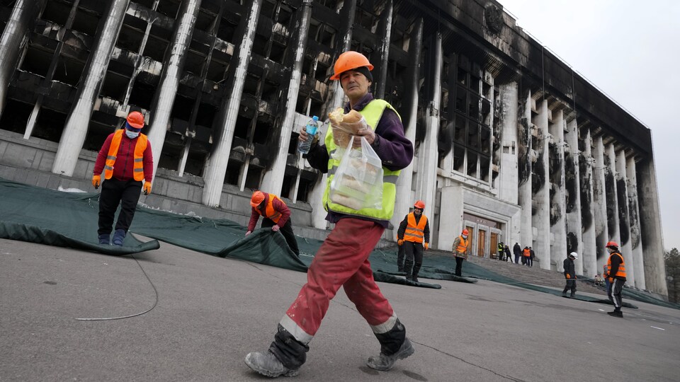 Un travailleur de la construction marche devant la façade calcinée d'un bâtiment en mangeant un sandwich.