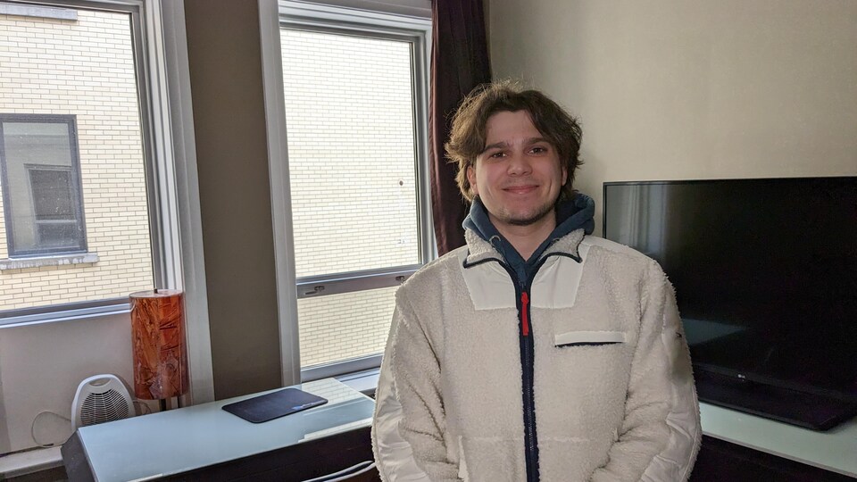 Rémi Covizzi dans sa chambre étudiante, une télévision se trouve derrière lui ainsi qu'un petit bureau. 