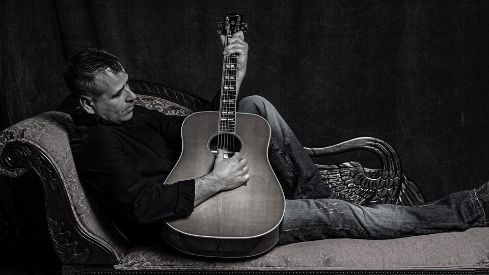 Hubert Cormier qui joue de la guitare confortablement installé sur un sofa.