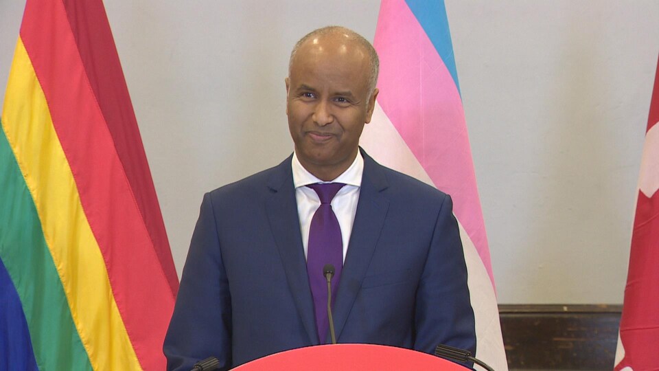 Le ministre Ahmed Hussen parle devant une tribune avec des drapeaux des communautés LGBTQ+ derrière lui.