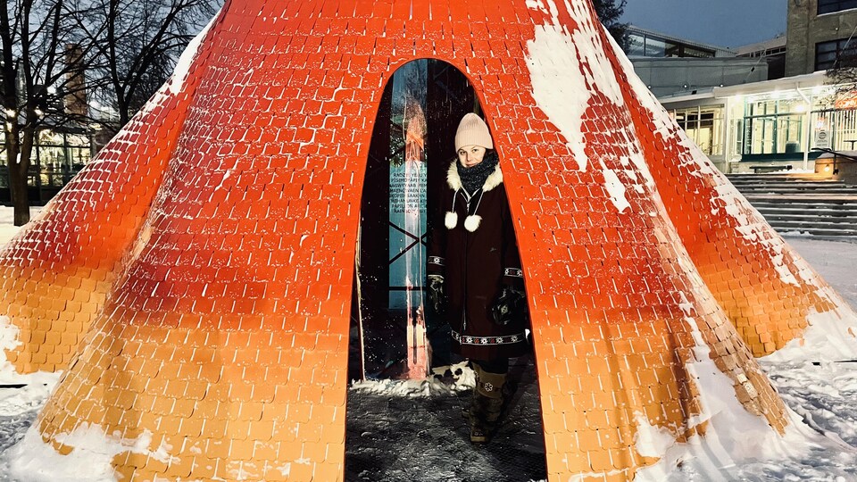 Une structure rouge et orange est déposée sur de la neige. Rachelle Lemieux est à l'intérieur avec son manteau et sa tuque.