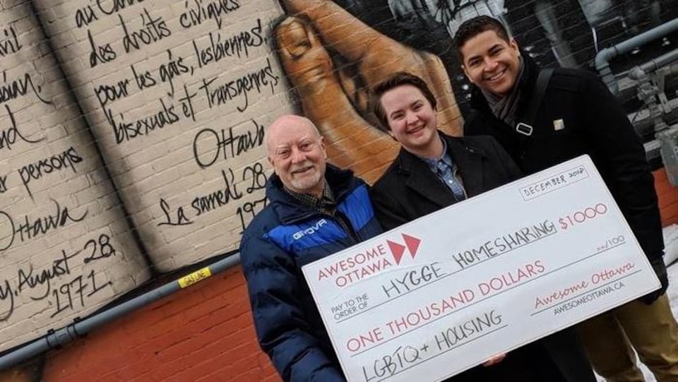 Deux hommes et une femme posent pour la caméra en tenant un gros ch`que devant une murale peinte sur le mur extérieur d'un bâtiment.