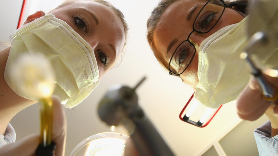 Deux femmes masquées apparaissent penchées au-dessus de l'appareil-photo avec, dans les mains, des outils de dentisterie.