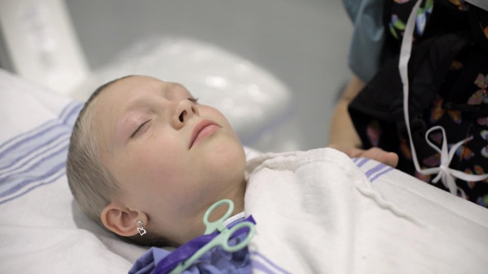 Une jeune fille aux cheveux très courts a les yeux fermés, allongée sur un lit d'hôpital.