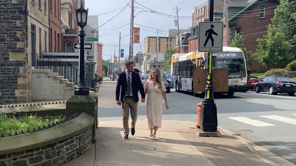 A man in dark clothes and a woman in long clothes walk hand in hand on the side of a street with cars and buses.