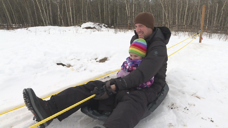 Ian Park tient sa fille dans ses bras sur une luge en hiver. Ils montent la pente de leur cour arrière grâce à une corde tirée vers le haut par un moteur.
