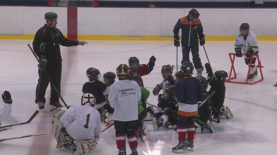 Un coach de hockey donne des consignes à un groupe de jeunes joueurs assis sur la glace.