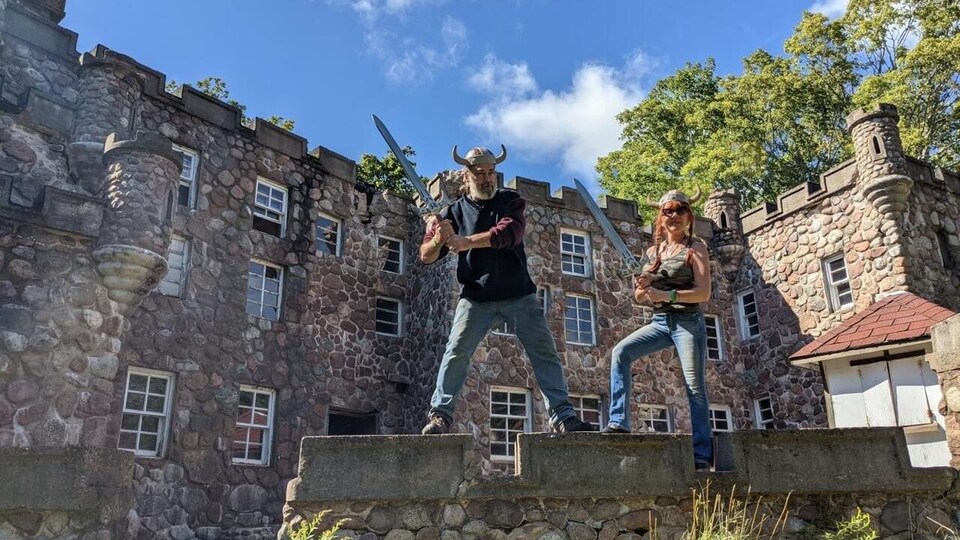 Un couple avec des fausses épées et des casques vikings font la pose devant un petit château de pierre.