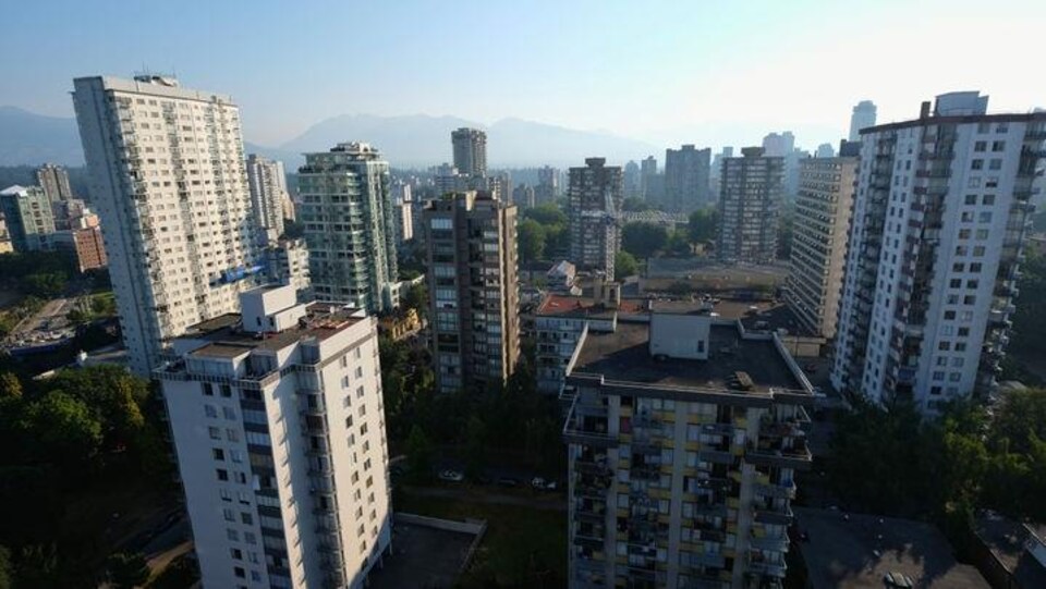 Point de vue de la ville de Vancouver depuis la fenêtre d'une tour à logements. On y voit plusieurs tours avec les montagnes en arrière plan.