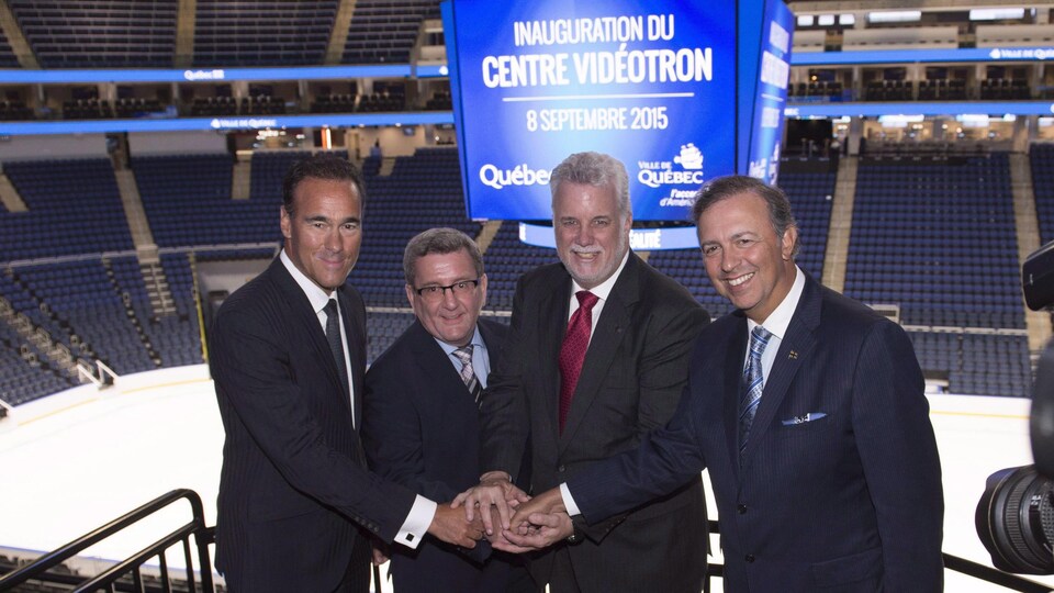 Les quatre hommes se tiennent la main en signe d'alliance. À l'arrière, on aperçoit la glace de la patinoire de hockey ainsi que les estrades vides.