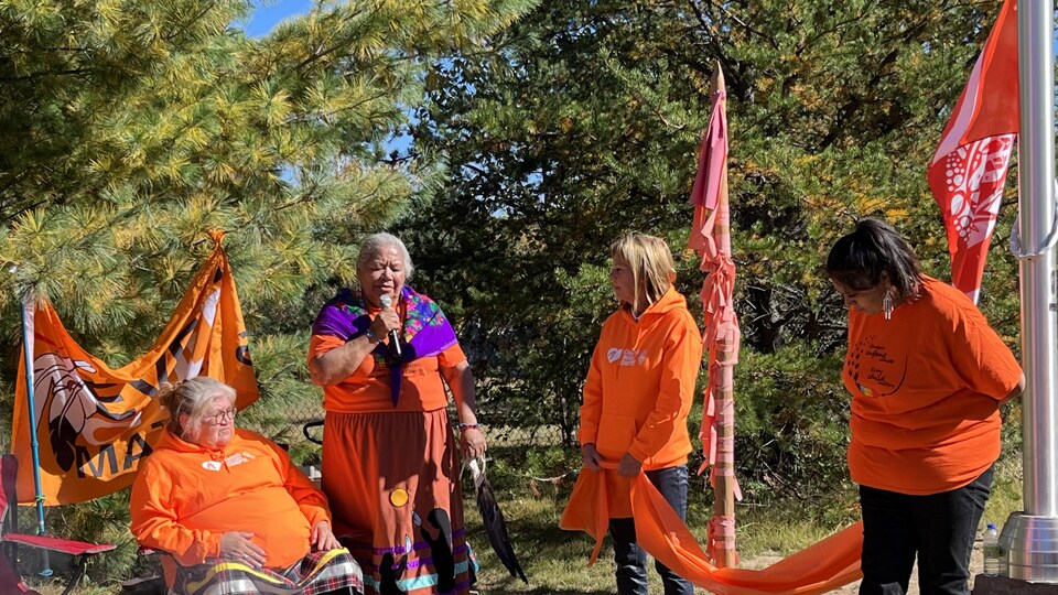Mary Coon parle au micro dans un parc. Elle est accompagnée de trois femmes toutes vêtues d'orange. 
