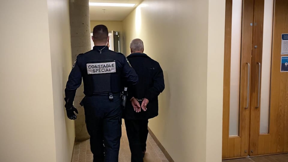 Claude Guillot, un homme âgé aux cheveux blancs, se fait escorter par un policier dans un couloir du palais de justice de Québec.