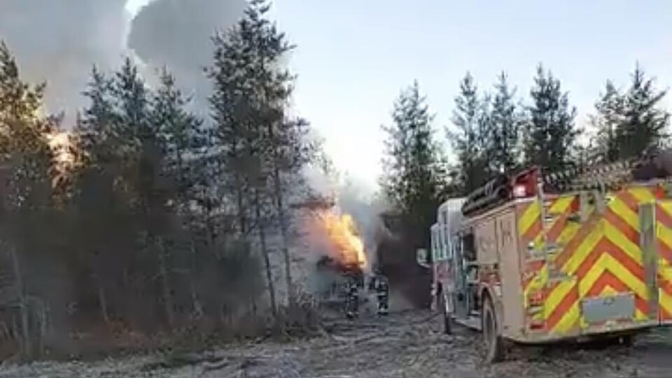Une photo du broyeur à chenilles en flammes, dans la forêt. Un camion de pompier est sur place. 