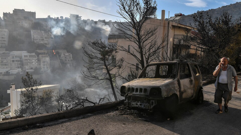 A man is walking next to a car that caught fire in the forest.