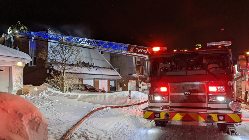 Un véhicule de pompiers devant un immeuble. Des pompiers s'affairent à éteindre le feu sur la toiture.