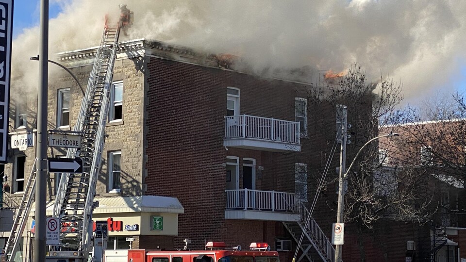 Des flammes et beaucoup de fumée sortent du toit d'un immeuble. Un pompier tente d'accéder au toit avec une échelle sur un camion.
