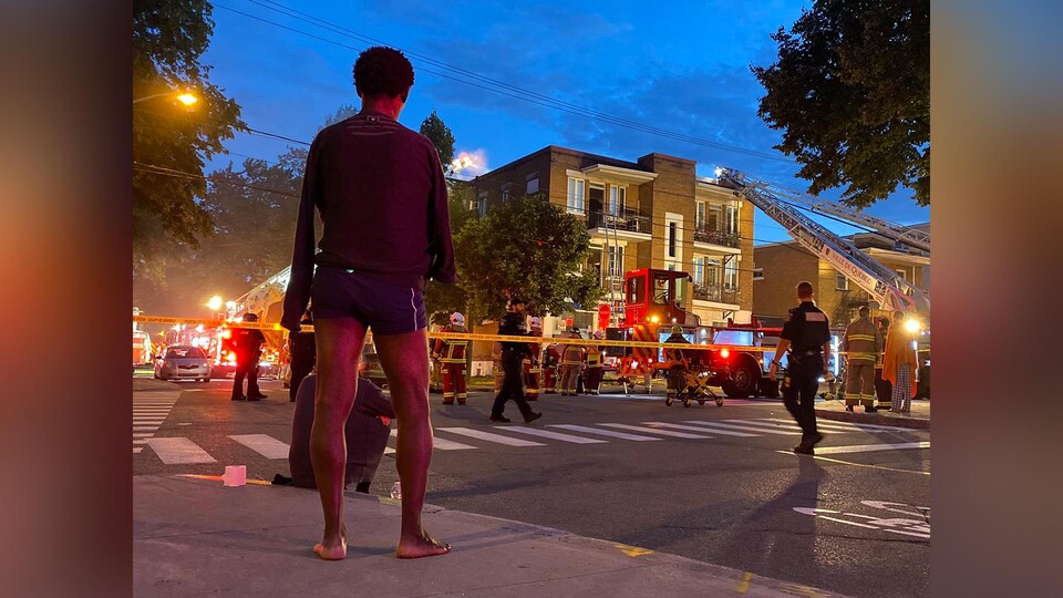 Un homme, de dos, regarde les pompiers à l'oeuvre. Il porte des boxers et se tient nu pied dans la rue.