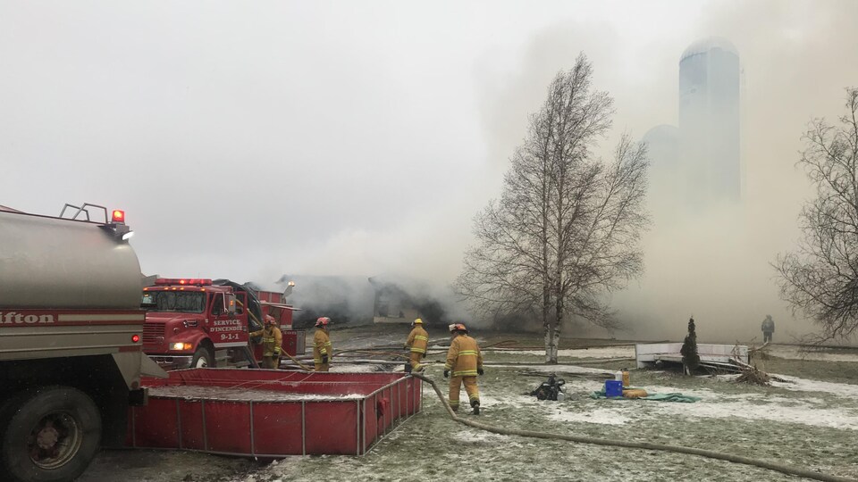 Plus d'une vingtaine de pompiers ont été appelés à combattre l'incendie sur le rang 5, à Saint-Malo. On voit ici les pompiers puiser de l'eau d'un bassin. Il y a énormément de fumée derrière eux.