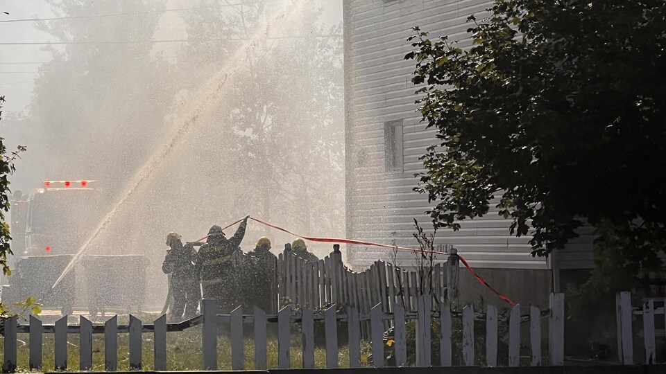 Pendant qu'une lance arrose l'immeuble d'eau, quelques pompiers s'affairent près de la base du bâtiment.