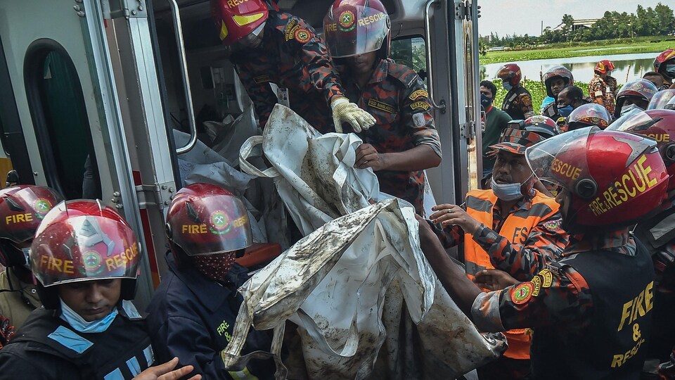 On voit des pompiers et des secouristes qui transportent un sac en tissu pour le mettre dans un véhicule.
