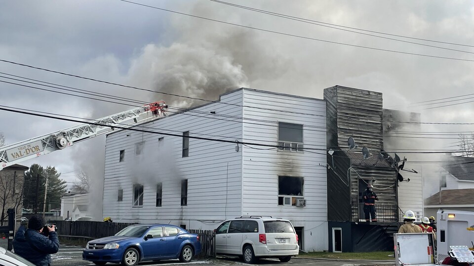 Des pompiers tentent d'éteindre le feu. Leur grue mène au toit où se dégage la fumée.
