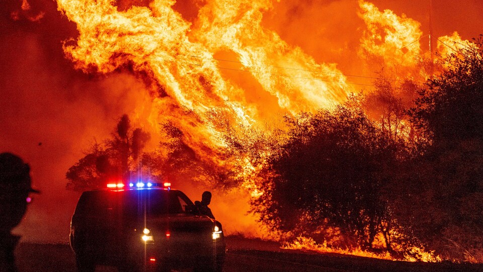 Un policier près de son véhicule de service regarde les flammes d'un incendie violent.