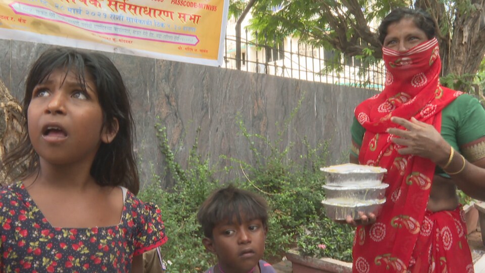 Uma mascarada segura refeições prontas e fala para a câmera, rodeada por duas crianças.