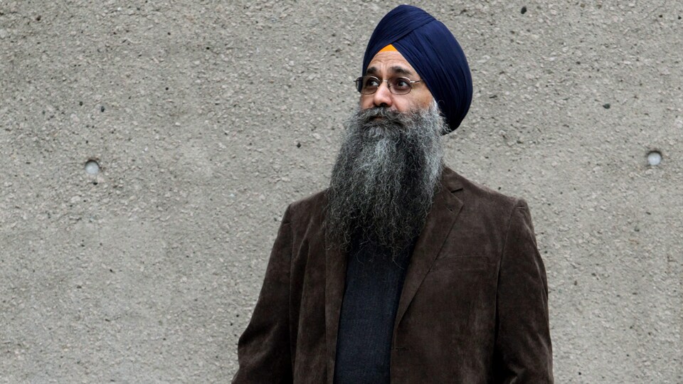 Un homme à la barbe longue portant un turban photographié devant un mur de béton.