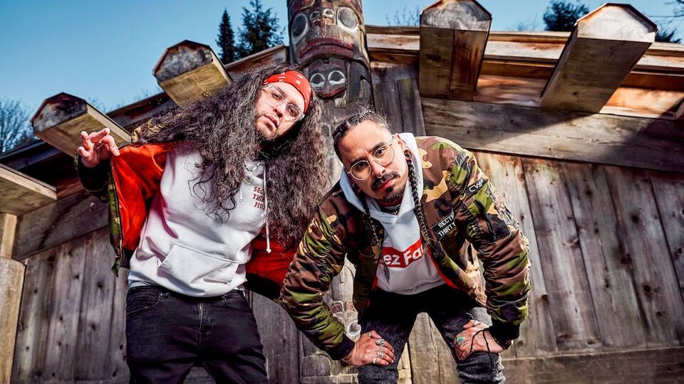 Deux hommes affichent un air sérieux devant un totem et une cabane en bois.
