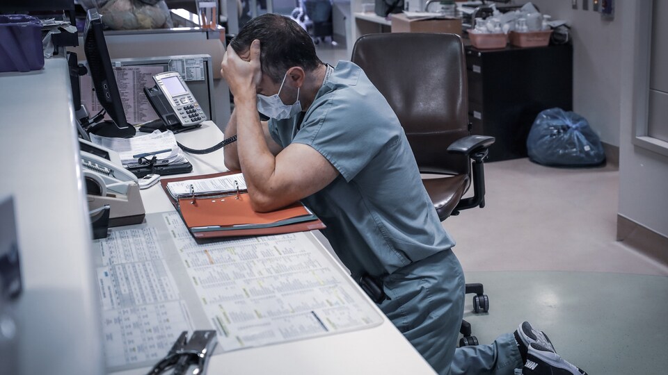 Un médecin en milieu hospitalier à genoux par terre et accoudé à une table. Une main sur son front et l'autre tient le combiné d'un téléphone.
